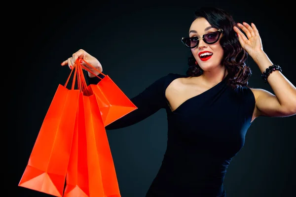 Black Friday Sale concept voor shop. Winkelen vrouw in zonnebril Holding rode tas geïsoleerd op donkere achtergrond. — Stockfoto