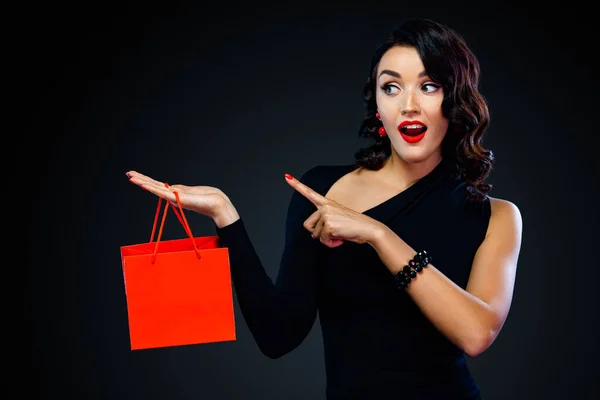 Fekete pénteki vásár koncepció a boltnak. Bevásárló lány kezében piros táska elszigetelt sötét háttérrel. A nő balra mutat a fénymásolón.. — Stock Fotó