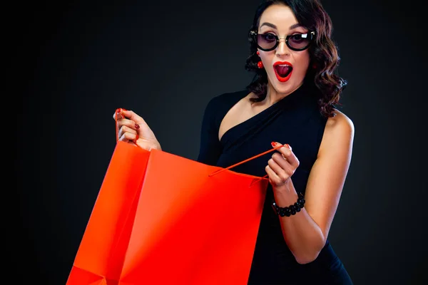 Koncepcja wyprzedaży Black Friday dla sklepu. Zaskoczona kobieta w okularach przeciwsłonecznych trzyma czerwoną torbę na ciemnym tle na zakupach. — Zdjęcie stockowe
