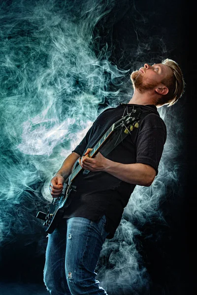 Handsome young musician man playing on guitar and singing in the smoke on stage or scene. — Stock Photo, Image