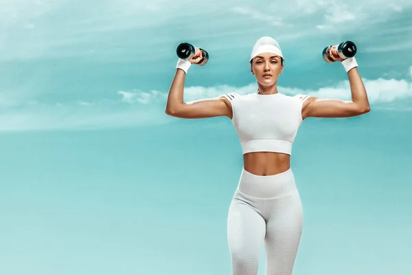 Sportieve en fitte vrouwelijke atleet doet training met halters op de achtergrond van de lucht. Het concept van een gezonde levensstijl en sport. Individuele sportrecreatie. — Stockfoto