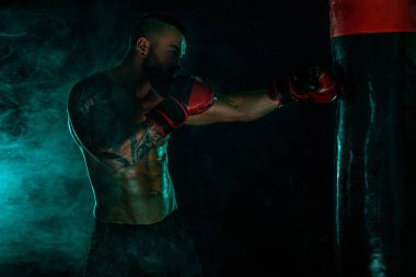 Boksör boks torbasıyla eldivenlerle dövüşüyor. Dövmeli bir sporcu, siyah arka planda dumanlı bir adam. Boşluğu Kopyala.