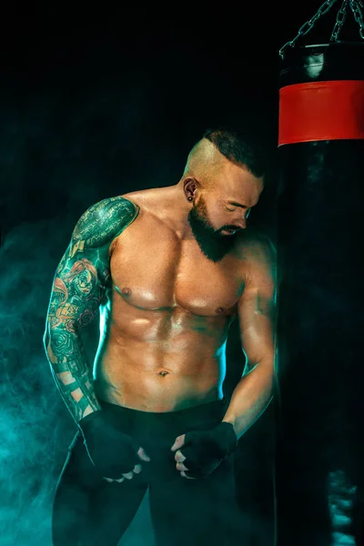 Boxeador luchando en guantes con saco de boxeo. Deportista con tatuajes, hombre aislado sobre fondo negro con humo. Copiar espacio . —  Fotos de Stock