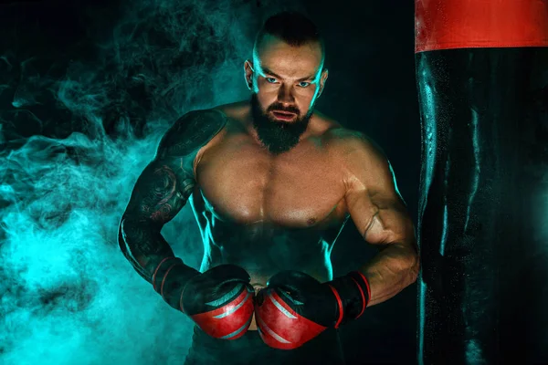 Boxeador luchando en guantes con saco de boxeo. Deportista con tatuajes, hombre aislado sobre fondo negro con humo. Copiar espacio . —  Fotos de Stock