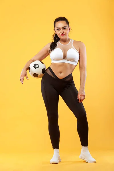 Talla más mujer en forma deportiva mujer en ropa deportiva con pelota de fútbol, atleta hace ejercicio de fitness sobre fondo amarillo. Motivación para las personas gordas . — Foto de Stock