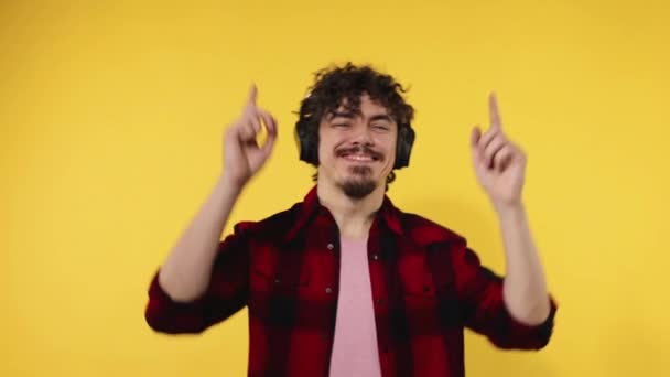 Handsome happy european man with beard in red shirt smiling and dancing isolated on yellow background. Guy in headphones listening to music. Lifestyle concept. Slow motion. — Stockvideo