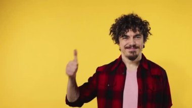 Man shows thumbs up sign with fingers. Closeup portrait of happy smiling guy with curly hair looking at camera isolated on yellow background. Slow motion.