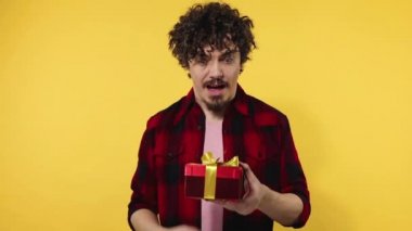 A man gives a red gift for Valentines Day lovers or International Womens Day. Birthday surprise. Handsome happy european man with beard in shirt smiling isolated on yellow background. Slow motion.