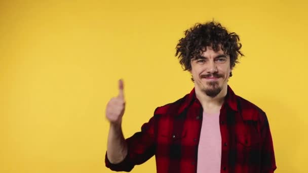 El hombre muestra el pulgar hacia arriba con los dedos. Retrato de primer plano del chico feliz sonriente con el pelo rizado mirando a la cámara aislada sobre fondo amarillo. Movimiento lento . — Vídeo de stock