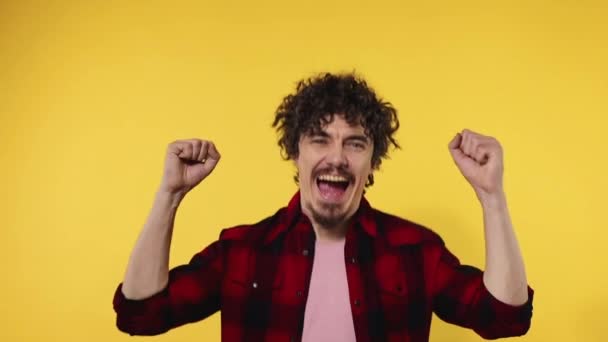 El hombre celebra la victoria del equipo. Retrato de primer plano del chico feliz sonriente con el pelo rizado mirando a la cámara aislada sobre fondo amarillo. Movimiento lento . — Vídeos de Stock