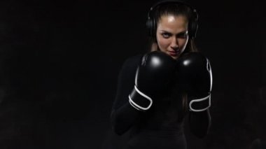 Spor salonunda boks antrenmanı yapan genç kadın sporcu boksör. Eldiven giyen, spor kıyafetler giyen ve kum torbasına vuran kız. Duman ile siyah arka plan üzerinde izole. Kopyalama Alanı.