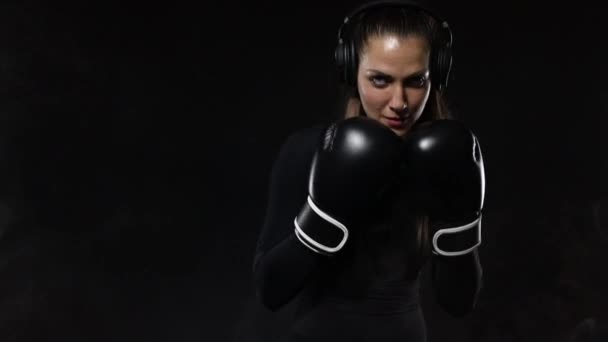 Young woman sportsman boxer doing boxing training at the gym. Girl wearing gloves, sportswear and hitting the punching bag. Isolated on black background with smoke. Copy Space. — Stock Video