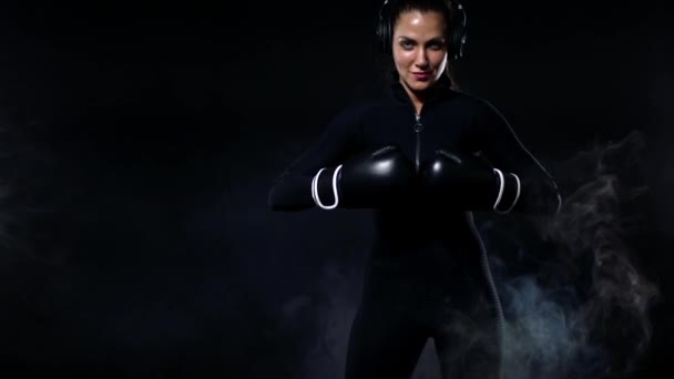 Mujer joven deportista boxeadora haciendo entrenamiento de boxeo en el gimnasio. Chica con guantes, ropa deportiva y golpear el saco de boxeo. Aislado sobre fondo negro con humo. Copiar espacio . — Vídeo de stock