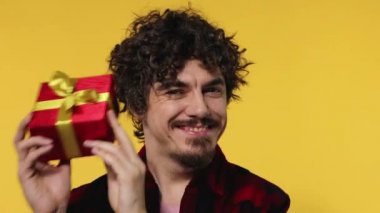 A man gives a red gift for Valentines Day or International Womens Day. Birthday surprise. Handsome happy european man with beard in shirt smiling isolated on yellow background.