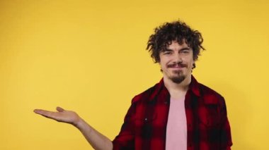 Man shows ok sign with fingers. Closeup portrait of happy smiling guy with curly hair looking at camera isolated on yellow background. Slow motion. Model pointing to looking left on copy space.