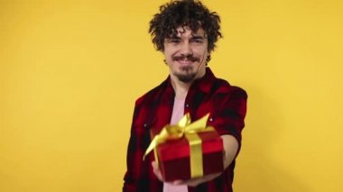 A man gives a red gift for Valentines Day lovers or International Womens Day. Birthday surprise. Handsome happy european man with beard in shirt smiling isolated on yellow background. Slow motion.