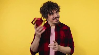 A man gives a red gift for Valentines Day lovers or International Womens Day. Birthday surprise. Handsome happy european man with beard in shirt smiling isolated on yellow background. Slow motion.