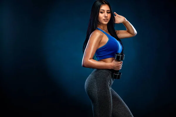Koyu arka planda dambılları olan güçlü atletik formalı bir kadın mavi spor giysisi giyiyor. Spor ve spor motivasyonu. — Stok fotoğraf