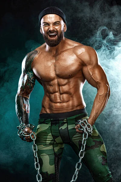 Athletischer Bodybuilder auf schwarzem Hintergrund mit Licht und Rauch. Männermode. Porträt eines brutalen bärtigen Mannes oben ohne mit Ketten. — Stockfoto
