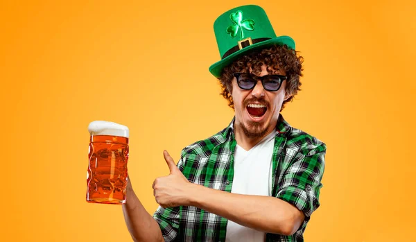 Il giorno di San Patrizio. Giovane uomo Oktoberfest che serve grande tazza di birra con bevanda isolata su sfondo giallo. Ragazzo mostrando pollice su segno con le dita . — Foto Stock