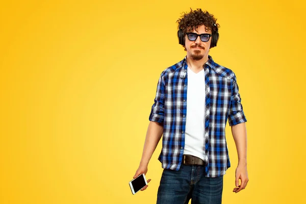 European man with curly hair in blue sunglasses with mobile phone or smarphone. Handsome smiled stylish hipster in plaid shirt posing over yellow background. — 图库照片