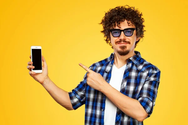 Europejczyk w niebieskich okularach przeciwsłonecznych z telefonem komórkowym lub smartfonem. Facet wskazujący na ekran. Przystojny, stylowy hipster w koszuli w kratę, pozujący na żółte tło. — Zdjęcie stockowe