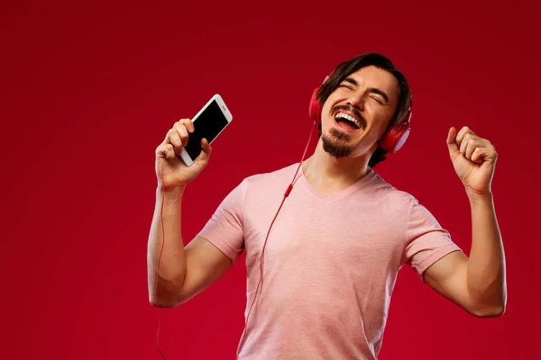 Przystojny, szczęśliwy Europejczyk z brodą w czerwonej koszulce, uśmiechnięty i tańczący na czerwonym tle. Facet w słuchawkach słuchający muzyki na smartfonie. Koncepcja stylu życia. — Zdjęcie stockowe
