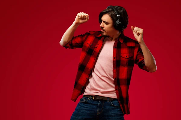 Guapo hombre europeo feliz con barba en camisa roja sonriendo y bailando aislado sobre fondo rojo. Un tipo con auriculares escuchando música. Concepto de estilo de vida . — Foto de Stock