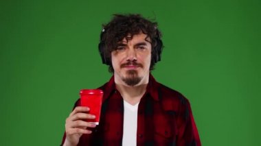 Kulaklıklı, kahve içip müzik dinleyen adam. Yeşil arka planda sakallı, gülümseyen ve dans eden yakışıklı, mutlu Avrupalı bir adam. Krom anahtar.