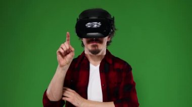 Kıvırcık saçlı, VR kulaklık kullanan ve yeşil arka planda izole edilmiş sanal gerçekliği deneyimleyen genç bir adam. Krom anahtar.