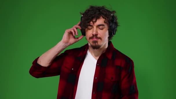 Guapo hombre europeo feliz con barba en camisa roja sonriendo y bailando aislado sobre fondo verde. Un tipo con auriculares escuchando música. Concepto de estilo de vida . — Vídeo de stock