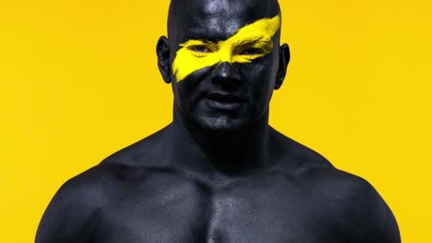 Man bodybuilder athlete with yellow color on face art and black body paint. Colorful portrait of the guy with bodyart. — Stock Video