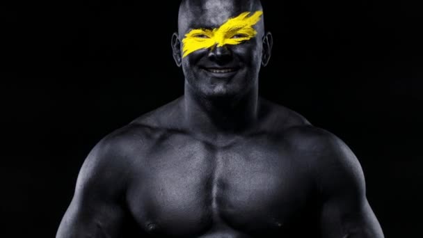 Man bodybuilder athlete with yellow color on face art and black body paint plays muscles on the chest. Colorful portrait of the guy with bodyart. — Stock Video