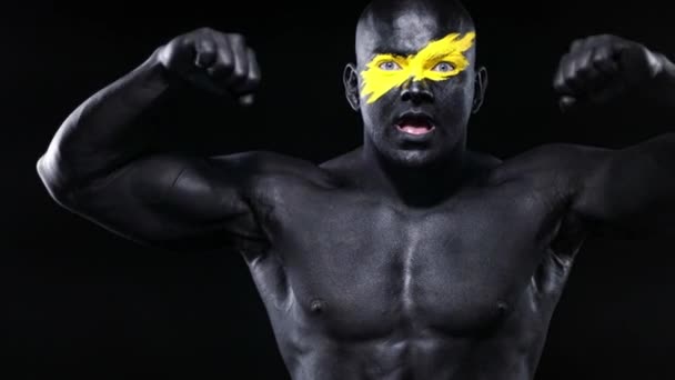 Uomo bodybuilder atleta con colore giallo sulla faccia arte e vernice corpo nero gioca bicipiti muscoli. Ritratto colorato del ragazzo con bodyart . — Video Stock