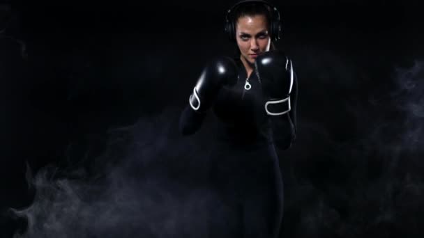 Młoda kobieta sportowca bokser robi szkolenia bokserskie na siłowni. Dziewczyna nosi rękawice, Odzież sportowa i uderzając worek wykrawania. Izolowane na czarnym tle z dymem. Kopiuj przestrzeń. — Wideo stockowe