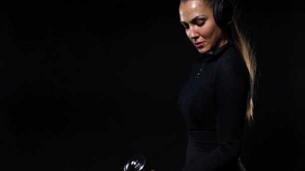 体育和健身概念。适合女运动员穿着运动服和耳机，用哑铃锻炼。被黑色背景隔离的女孩。慢动作, — 图库视频影像