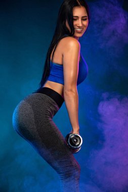 Koyu arka planda dambılları olan güçlü atletik formalı bir kadın mavi spor giysisi giyiyor. Spor ve spor motivasyonu.