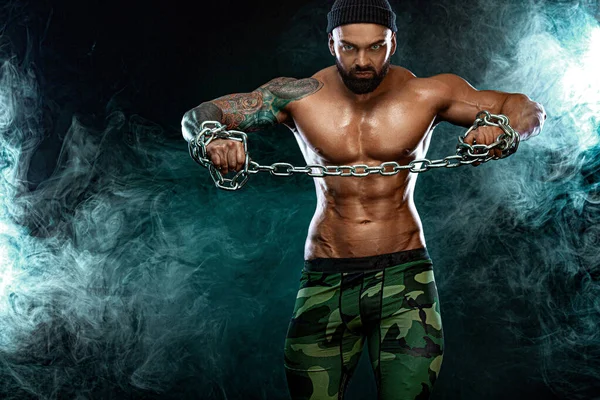 Atleta bodybuilder su sfondo nero con luci e fumo. Moda uomo. Ritratto di un brutale uomo barbuto in topless con catene . — Foto Stock