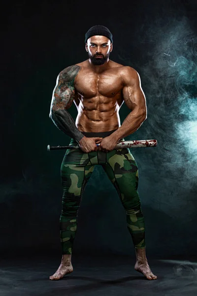 Forte e apto fisiculturista homem com bastão de beisebol mostra músculos abdominais sob uma camiseta. Um atleta musculoso desportivo. Conceito de desporto e fitness. Moda masculina . — Fotografia de Stock
