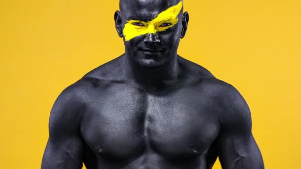 Man bodybuilder athlete with yellow color on face art and black body paint plays muscles on the chest. Colorful portrait of the guy with bodyart. — Stock Video