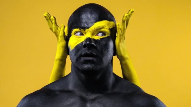 Homme avec des mains féminines sur le corps. Athlète bodybuilder avec l'art du visage jaune et la peinture de corps noire. Portrait coloré du gars avec bodyart . — Video