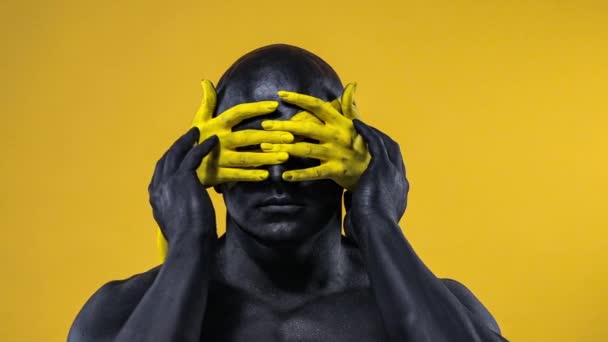 Man with female hands on the body. Bodybuilder athlete with yellow face art and black body paint. Colorful portrait of the guy with bodyart. — Stock Video