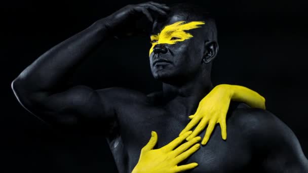 Homme avec des mains féminines sur le corps. Athlète bodybuilder avec l'art du visage jaune et la peinture de corps noire. Portrait coloré du gars avec bodyart . — Video