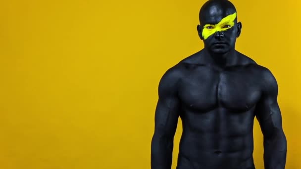 Man bodybuilder athlete with yellow color on face art and black body paint. Colorful portrait of the guy with bodyart. Slow motion. — Stock Video