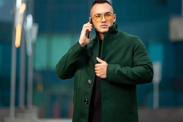 Jovem homem de negócios em casaco verde no centro de negócios com smartphone. Outono ou primavera homens moda olhar . — Fotografia de Stock