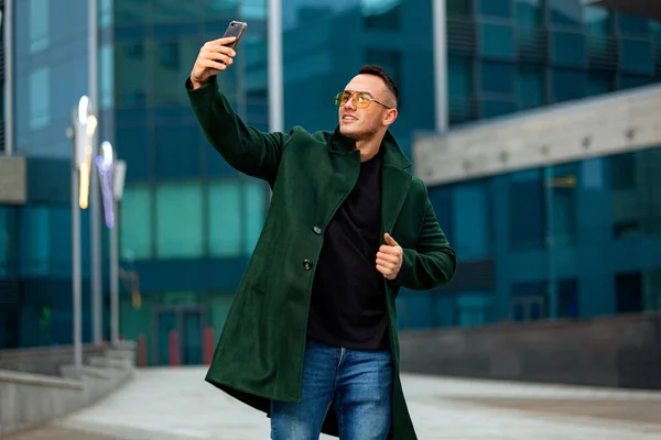 Jovem homem de negócios em casaco verde no centro de negócios com smartphone fazer selfie — Fotografia de Stock