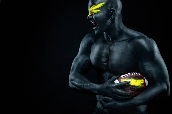 Fan de football américain avec ballon sur fond noir. Fitness et motivation sportive. Forte forme et gars athlétique dans la peinture du corps comme un super héros . — Photo