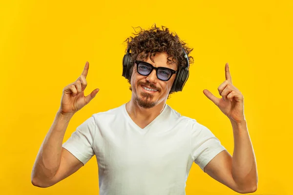 Porträt eines glücklichen bärtigen Mannes mit lockigem Haar, der mit dem Finger auf einen Bildschirm vor gelbem Hintergrund zeigt. Kerl mit blauer Sonnenbrille und Kopfhörer greift großartige Idee auf. — Stockfoto