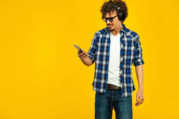 European man with curly hair in blue sunglasses with mobile phone or smarphone. Handsome smiled stylish hipster in plaid shirt posing over yellow background. — ストック写真