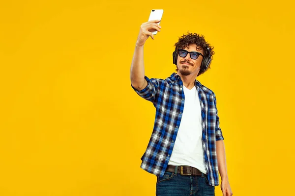 European man with curly hair in blue sunglasses with mobile phone or smarphone. Handsome smiled stylish hipster in plaid shirt posing over yellow background. — 图库照片